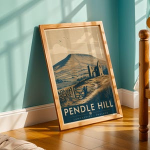 May include: A framed print featuring a vintage-style illustration of Pendle Hill in Lancashire, England. The artwork depicts a mountain landscape with a stone wall, gate, and ruins. The print has a wooden frame and is leaning against a wall.