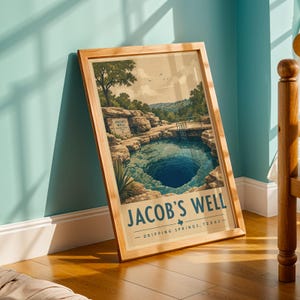 May include: A framed art print featuring a landscape illustration of Jacob's Well in Dripping Springs, Texas. The image depicts a deep blue natural spring surrounded by rocky cliffs, trees, and a small ladder, with the text Jacob's Well at the bottom.