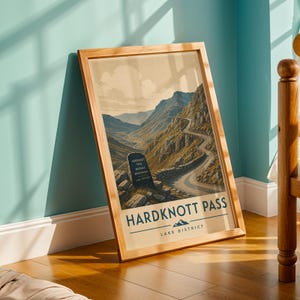 Op de afbeelding: Een ingelijste print met een vintage reisposterontwerp. De poster toont een kronkelende weg door een bergpas, met de tekst "Hardknott Pass Lake District" onderaan. De lijst is gemaakt van lichtgekleurd hout.