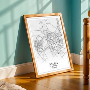Può includere: Una stampa incorniciata di una mappa di Medina, Arabia Saudita, in bianco e nero. La mappa è dettagliata, mostrando strade e la disposizione della città. La cornice è in legno chiaro. Il testo in basso recita "Medina - Saudi Arabia - City of Light."