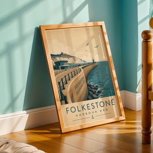 May include: A framed print of a vintage travel poster for Folkestone Harbour Arm. The image shows a coastal scene with a pier, lighthouse, and the words "Folkestone Harbour Arm". The frame is made of light wood and is leaning against a wall.