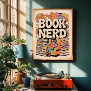 以下が含まれることがあります： レトロなフォントで「BOOK NERD」という言葉が書かれた額入りのポスター。本に囲まれた椅子で読書をする人の漫画が描かれています。ポスターは金色のフレームで、ティール色の壁に掛けられています。