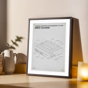 Peut inclure: Impression encadrée du système de divertissement Nintendo original (NES). Le schéma montre les composants et les étiquettes de la console. L'impression est dans un cadre noir, posée sur une surface en bois, avec une cruche blanche et une bougie en arrière-plan.