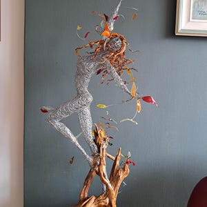Peut inclure: Sculpture en fil de fer représentant une danseuse aux cheveux flottants et aux bras tendus. La figurine est ornée d'éléments en forme de feuilles colorées. La sculpture est fixée sur une base en bois, créant une œuvre d'art unique.