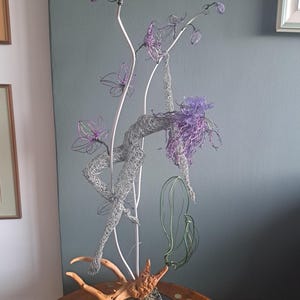 May include: A wire sculpture of a figure in a graceful pose, with flowing purple hair and delicate wire flowers. The sculpture is mounted on a base with a piece of driftwood. The figure is made of silver wire.