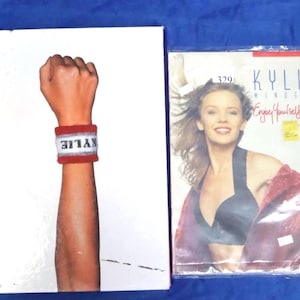 以下が含まれることがあります： 2つのアイテムが表示されています。白いアルバムカバーには「KYLIE」と書かれた赤と白のリストバンドをつけた拳があり、カイリー・ミノーグの「Enjoy Yourself Tour」のアルバムカバーがあります。