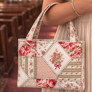 May include: A patchwork tote bag with a floral pattern in shades of red, pink, and green. The bag features a rectangular shape with two handles and is held by a person. The background shows a church interior.