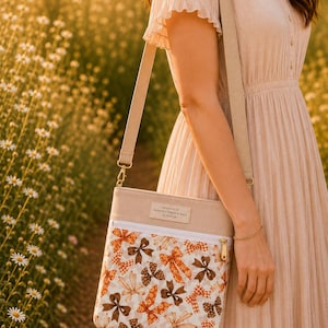 May include: A beige crossbody bag featuring a white zippered front pocket with a repeating pattern of orange and brown bows. The bag has a wide beige adjustable strap and a rectangular label at the top that reads HANDMADE BY HEAVENLY THREADS BAGS.