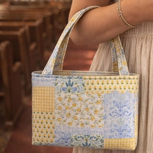 Op de afbeelding: Een patchwork tote bag met een rechthoekige vorm en twee handvatten. De tas heeft een mix van bloemen-, ruit- en damastpatronen in geel, blauw en wit. De tas wordt door een persoon vastgehouden.