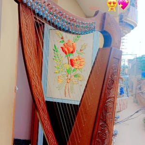 May include: A detailed wooden harp, featuring decorative carvings and a painted floral panel. The harp has numerous strings and tuning pegs. The soundboard displays a bouquet of orange roses. The instrument has a rich, brown finish.