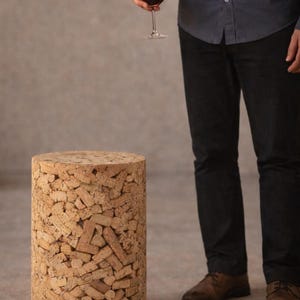 Pode incluir: Um banco cilíndrico feito de pedaços de cortiça. O banco é castanho claro e tem uma superfície texturizada. Uma pessoa segura um copo de vinho tinto perto do banco. O banco é uma peça de mobiliário única.