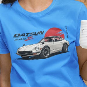 Może przedstawiać: Jasnoniebieski t-shirt z grafiką przedstawiającą biały samochód sportowy Datsun 240Z. Wzór zawiera czarno-czerwony napis DATSUN 240Z, duże czerwone koło w tle oraz pionowo ułożone japońskie znaki po prawej stronie koszulki.