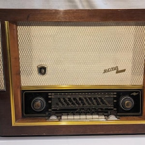 Puede incluir: Radio de madera vintage con cubierta de altavoz color crema y detalles dorados. La radio tiene un panel de control negro con diales y un dial de sintonización. La marca "REMA" es visible.