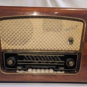 Puede incluir: Radio de madera vintage con rejilla de altavoz dorada y panel de control negro. La radio tiene un acabado marrón oscuro brillante y un emblema decorativo sobre el altavoz. Se ven perillas y botones en la parte delantera.