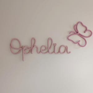 May include: A light pink, knitted wall decoration spelling the name "Ophelia" in cursive script, with a matching butterfly. The name and butterfly are mounted on a plain, off-white wall, creating a delicate, handmade aesthetic.