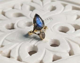 Può includere: Un anello color oro con due pietre preziose a forma di goccia. La pietra più grande ha sfumature blu e grigie, mentre quella più piccola è di un grigio più chiaro. Il design dell'anello include una fascia delicata e un'incastonatura intricata. Il testo "ENDLESS EMERAL" è visibile.