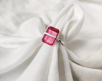 May include: A silver ring featuring a rectangular, faceted, bright pink gemstone. The gemstone is set horizontally, bisected by a silver bar. The ring is displayed on a white, textured fabric, creating a contrast between the cool metal and the warm pink hue.