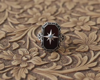 May include: A silver ring with a rectangular, dark red stone. A light blue starburst design is set in the center. The ring has floral accents on the sides and is set against a carved, floral background.