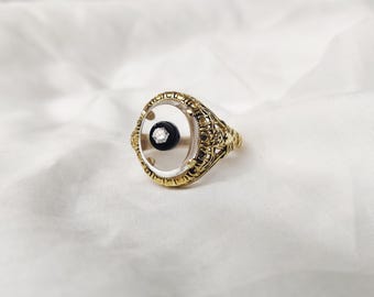 May include: A gold ring with an oval, clear centre. The centre has a black stone with a small diamond. The ring has an ornate, filigree design around the edges, giving it a vintage look.