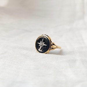 May include: A gold ring with a black oval centre stone featuring a white starburst design with a small diamond in the centre. The ring has a detailed, ornate band.