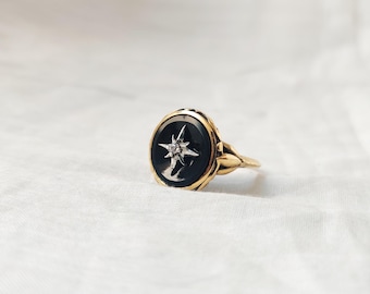 May include: A gold ring with a black oval center stone featuring a white starburst design with a small diamond in the center. The ring has a detailed, ornate band.