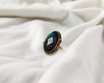 May include: A gold ring with an oval, dark green and black stone. The stone has a diamond-shaped, clear gem in the center. The ring has a detailed gold band and setting, and is set against a white fabric background.