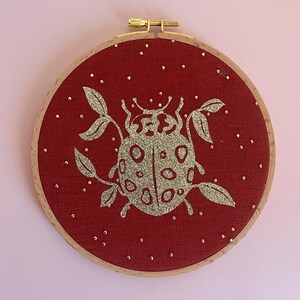 Peut inclure: Un cercle à broder rond avec un tissu rouge. Il présente un motif de coccinelle doré à paillettes, des feuilles et de petits points dorés. Le cercle en bois a un fermoir doré.