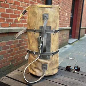 Könnte beinhalten: Eine zylindrische, braune Leder-Duffel-Tasche mit dunkelbraunen Lederriemen und Akzenten. Die Tasche hat einen Kordelzugverschluss oben und einen langen, weißen Riemen. Eine Sonnenbrille im Aviator-Stil liegt auf einer Holzoberfläche.