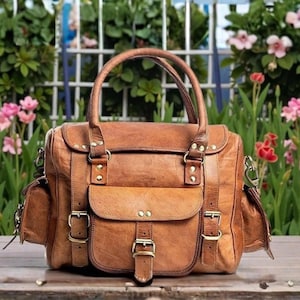 Puede incluir: Un bolso de cuero marrón con asa superior, bolsillo delantero y bolsillos laterales. El bolso tiene herrajes dorados, incluyendo hebillas y remaches. El bolso está sobre una superficie de madera con un fondo de flores y vegetación.