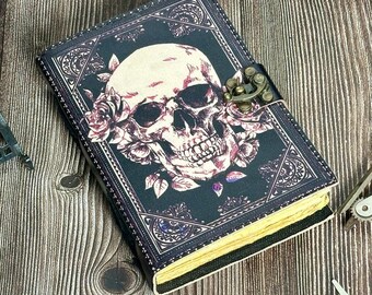 May include: A dark-colored journal with a skull and floral design on the cover. The skull is white and pink, surrounded by pink roses and green leaves. The journal has a metal clasp and yellowed pages.