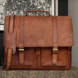 Puede incluir: Un maletín de cuero marrón con asa superior y bandolera. La bolsa tiene dos bolsillos delanteros y se asegura con dos cierres de hebilla. La correa tiene un reposahombros acolchado. El maletín está sobre una superficie estampada.