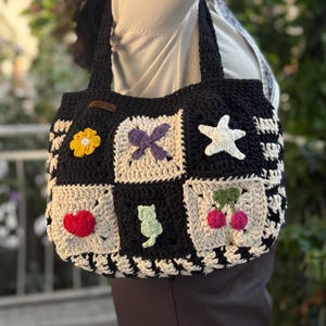 May include: A black and white crochet tote bag with a patchwork design. The bag features squares with various appliqués, including a yellow flower, a purple bow, a white star, a red heart, a green cat, and cherries. The bag has black handles.