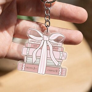 Op de afbeelding: Een heldere acryl sleutelhanger met een stapel roze boeken vastgebonden met een roze strik. De boeken hebben titels als "Enemies" en "Proximity". De sleutelhanger is bevestigd aan een zilveren metalen ring.