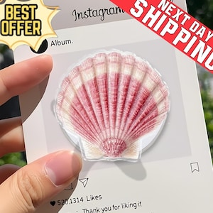 May include: A seashell-shaped phone grip with red and white details. The grip is attached to a white card with Instagram-themed graphics and the text "Album." and "520.1314 Likes." A "Best Offer" sticker and a "Next Day Shipping" banner are also visible.