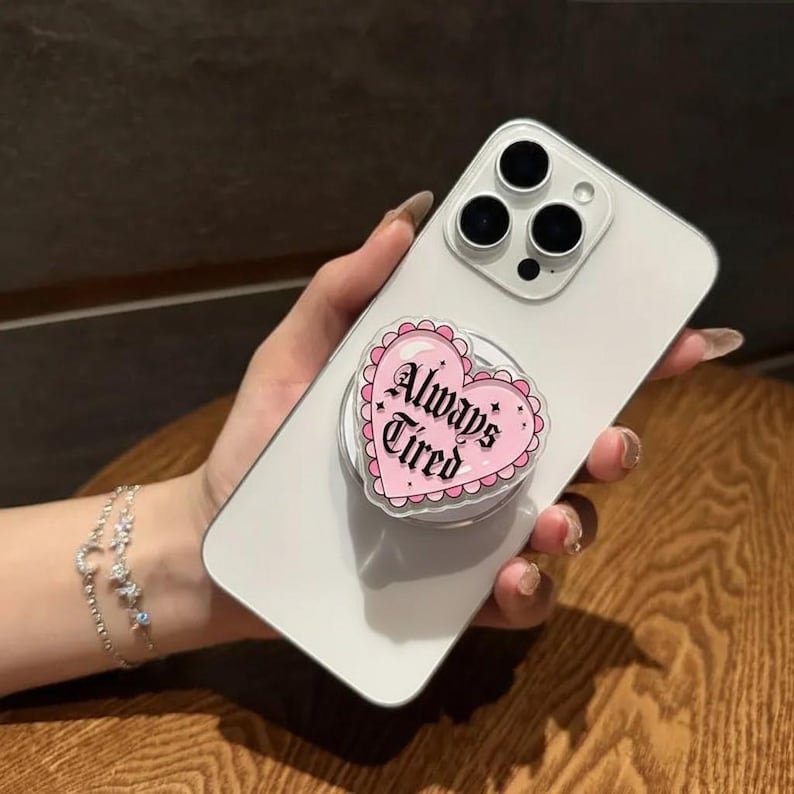 May include: A white smartphone with a pink heart-shaped phone grip attached. The phone grip has the words "Always Tired" in a decorative font. The grip is surrounded by a scalloped edge.