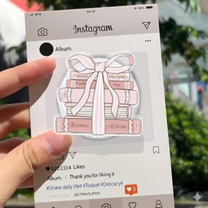 May include: A clear acrylic sticker showing a stack of pink books tied with a pink bow. The books have titles such as "Enemies to Lovers" and "Proximity." The sticker is set against a light grey Instagram-themed background.