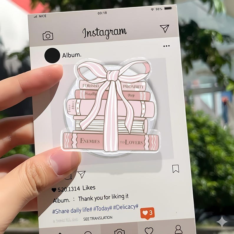 May include: A clear acrylic sticker showing a stack of pink books tied with a pink bow. The books have titles such as "Enemies to Lovers" and "Proximity." The sticker is set against a light grey Instagram-themed background.