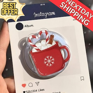 May include: A clear phone grip featuring a red mug design filled with marshmallows, candy canes, and a cinnamon stick. The mug has a snowflake design. The grip is set against a background that mimics an Instagram post, with the text "Instagram" visible.