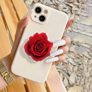 Könnte beinhalten: Cremefarbene Smartphone-Hülle mit einem leuchtend roten Rosenmuster. Die Hülle hat ein Dual-Kamera-System. Die Rose blüht voll, mit detaillierten Blütenblättern.