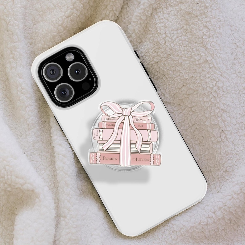 May include: White phone case featuring a stack of pink books with a pink bow illustration. The books are labelled with titles such as "Enemies to Lovers". The phone case has a black camera module.