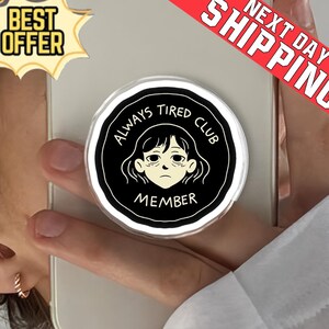 May include: A round phone grip with a black and white design. The graphic features a tired-looking face with the words "ALWAYS TIRED CLUB MEMBER" in a circular pattern. The grip is held on a phone.
