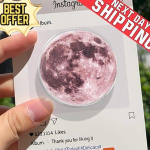 Puede incluir: Un soporte redondo para teléfono con un diseño de luna rosa. El soporte se presenta sobre un fondo blanco con gráficos de estilo redes sociales. La imagen incluye el texto "BEST OFFER" y "NEXT DAY SHIPPING".