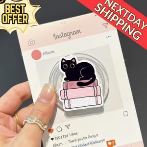 Può includere: Un adesivo di un gatto nero è appoggiato su una pila di libri rosa, su uno sfondo di un post di Instagram simulato. L'adesivo è su un supporto per telefono trasparente. Sono visibili anche le parole "BEST OFFER" e "NEXTDAY SHIPPING".