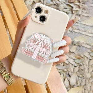 May include: A cream phone case with a pop-up grip showing a stack of pink books tied with a pink bow. The books have titles like "Enemies to Lovers" and "Proximity". A gold watch is visible on the wrist.