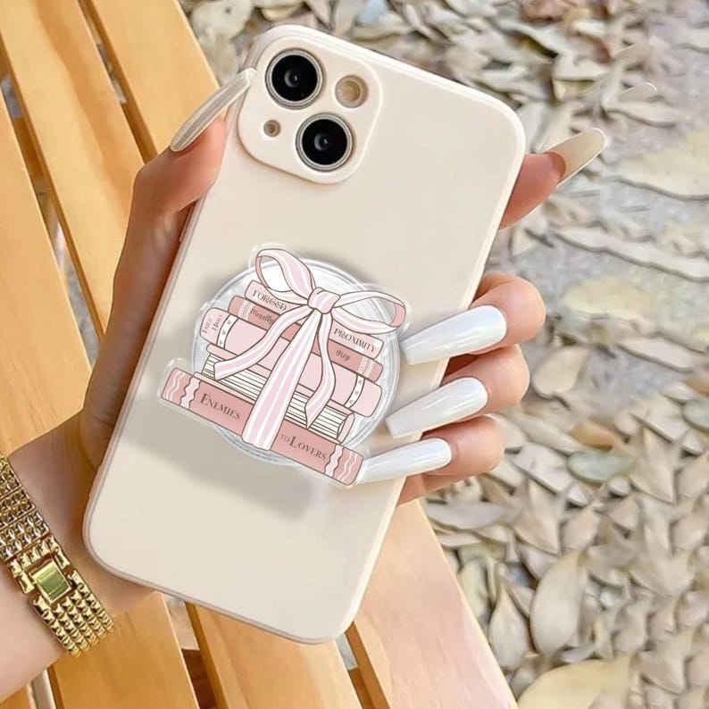 May include: A cream phone case with a pop-up grip showing a stack of pink books tied with a pink bow. The books have titles like "Enemies to Lovers" and "Proximity". A gold watch is visible on the wrist.
