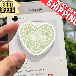 Op de afbeelding: Een lichtgroene hartvormige telefoonhouder met een bloemen- en boekenontwerp. De houder wordt weergegeven op een witte kaart met een Instagram-achtige lay-out. De kaart bevat de tekst "Album" en "520.1314 Likes".