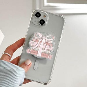 May include: Clear phone case featuring a stack of pink books and a pink bow design. The books have the words "Enemies to Lovers", "Proximity", and "Forced" printed on them. The phone case is held by a person.