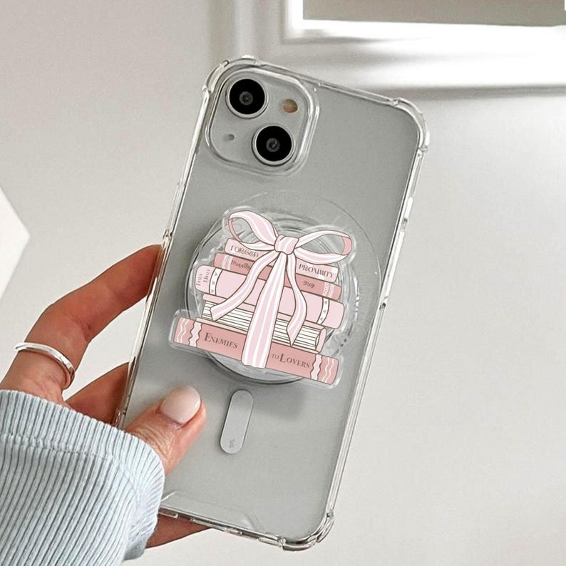 May include: Clear phone case featuring a stack of pink books and a pink bow design. The books have the words "Enemies to Lovers", "Proximity", and "Forced" printed on them. The phone case is held by a person.