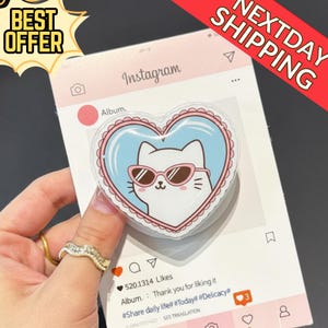 May include: A heart-shaped phone grip featuring a cartoon cat wearing sunglasses, set against a light blue background with a pink border. The grip is on a card designed to look like an Instagram post. The card has the text "Instagram" and "Album."