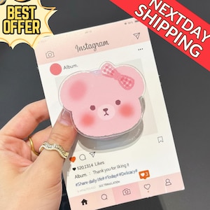 May include: A pink bear-shaped phone grip with a gingham bow, set against a pink Instagram-themed background. The image includes the text "BEST OFFER" and "NEXTDAY SHIPPING". The Instagram post shows a like count and a caption.